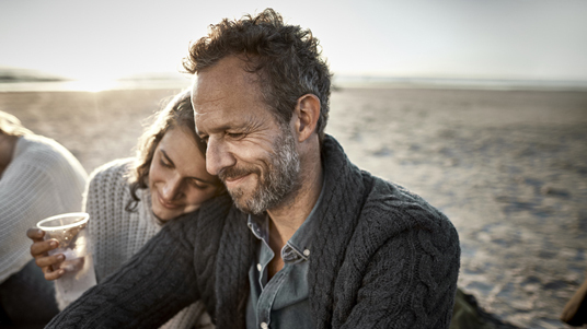 Pareja sentada en la playa sonriendo, con amanecer de fondo. La mujer apoya la cabeza en el hombro del hombre