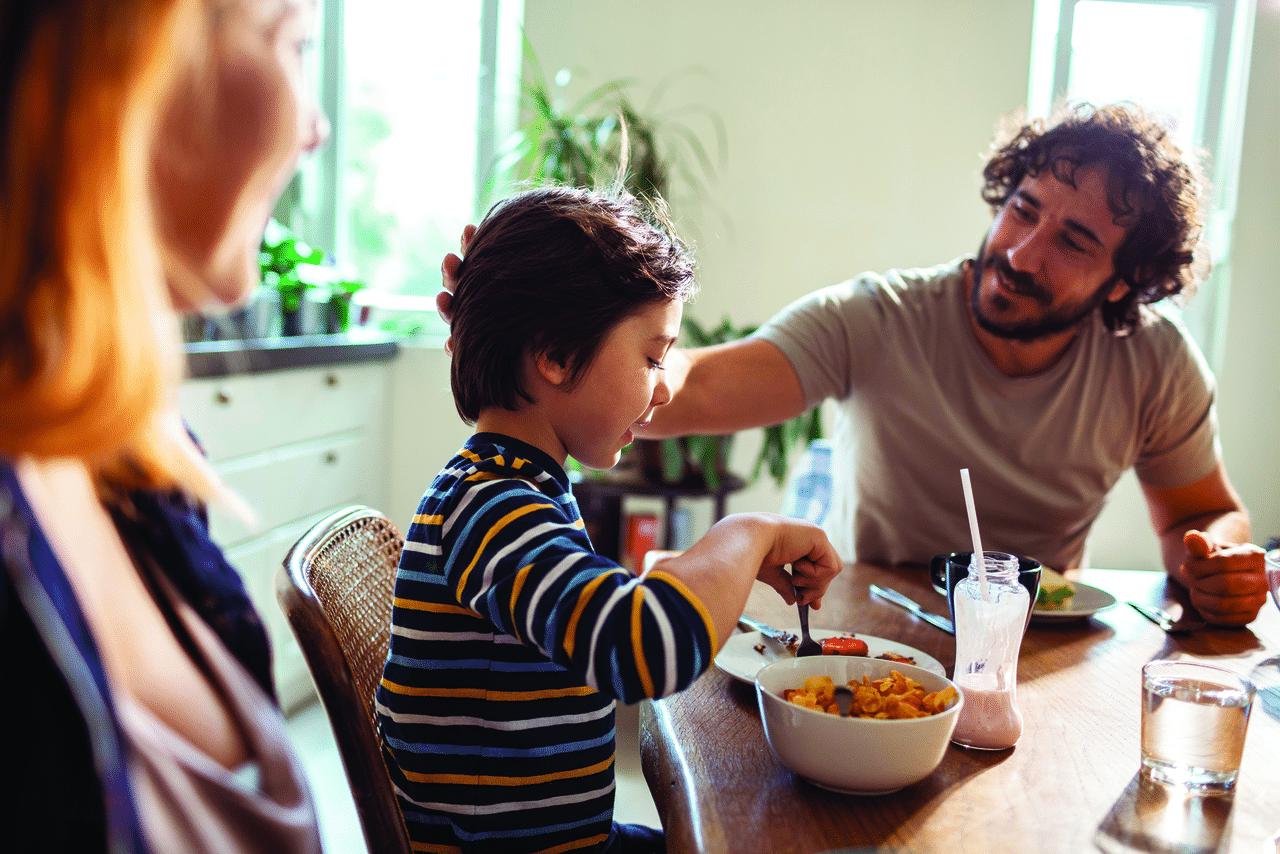 Niño comiendo sentado a la mesa, mientras su padre lo observa alentadoramente